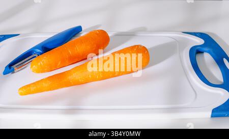 Two peeled carrots and a vegetable peeler placed on a white and blue cutting board. Stock Photo
