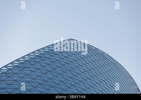 Modern architectural structure features sleek, curved surface with triangular glass panel grid. Clear blue sky backdrop emphasizes futuristic design. Stock Photo