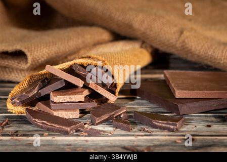 Pieces of artisanal dark and milk chocolate inside a small burlap sack, on a rustic wooden table next to chocolate bars. Stock Photo