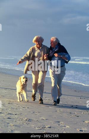 Senior man & woman running on the beach with their dog. Stock Photo