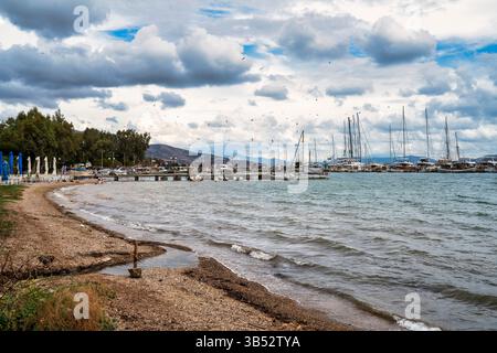 A peaceful marina in Nea Peramos, Greece, featuring docked sailboats, a sandy beach, and flocks of birds flying under dramatic clouds. Stock Photo