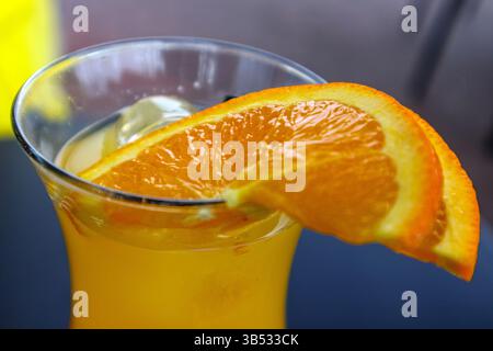 A close-up shows a glass of an amber-colored drink, possibly orange juice or a cocktail, with ice cubes and two orange slices on the rim of the glass. Stock Photo