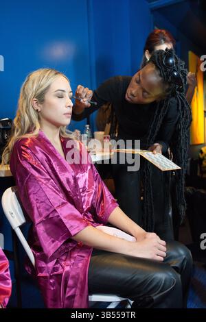 A young woman in a pink robe sits while a makeup artist applies eye makeup. The setting appears to be a backstage area. Stock Photo