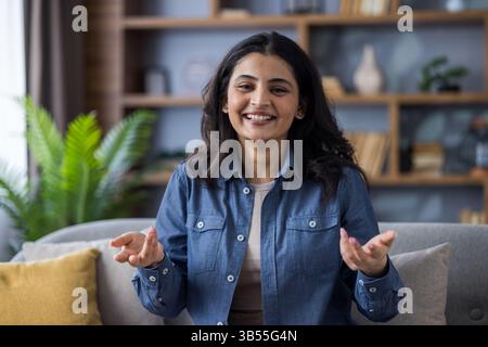 Close-up portrait of a young Indian woman in a denim shirt sitting on a sofa at home in front of the camera and talking while gesturing with her hands. Stock Photo