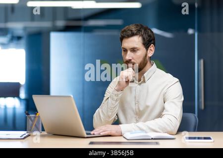 A young serious business man is sitting in the office at a desk and is concentrating on working on a laptop. Stock Photo