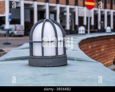 A close-up of a decorative light fixture on a stone wall, with a blurred urban background featuring buildings and vehicles. The light is enclosed in a Stock Photo