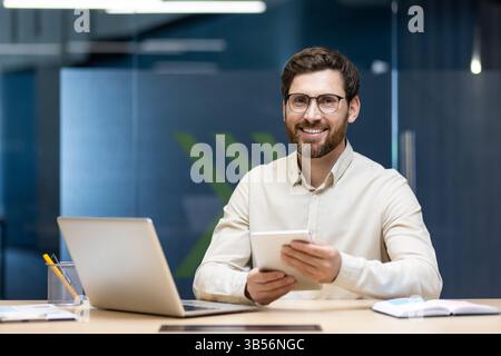 Portrait of a smiling and successful young businessman working in the office, sitting at a desk, holding a tablet and looking at the camera. Stock Photo