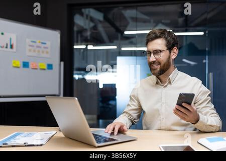 Smiling young man businessman, developer works in office at laptop on whiteboard with stickers, types on keyboard, holds mobile phone in hand. Stock Photo