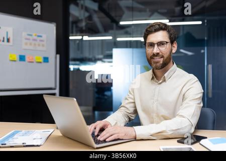 Portrait of a smiling young man sitting in the office behind a laptop and looking smugly at the camera. Stock Photo