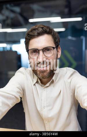 Vertical close-up photo of a young man in glasses and a shirt who is in the office and prays to the online camera he is holding in his hands. Stock Photo