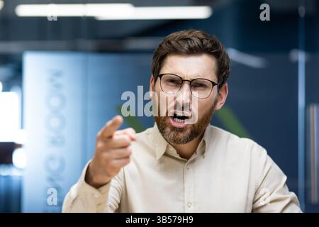 Close-up photo of a young angry businessman sitting in an office with a desk in front of the camera and talking angrily on a video call while pointing his finger at the camera. Stock Photo