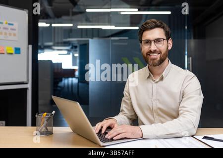 Portrait of a young successful man in glasses and a light shirt sitting in the office at the table, working on a laptop, smiling and looking at the camera. Stock Photo