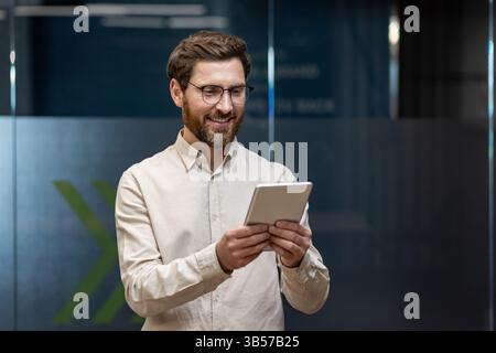 Smiling young male businessman standing in office wearing glasses and tie, holding and using tablet. Stock Photo