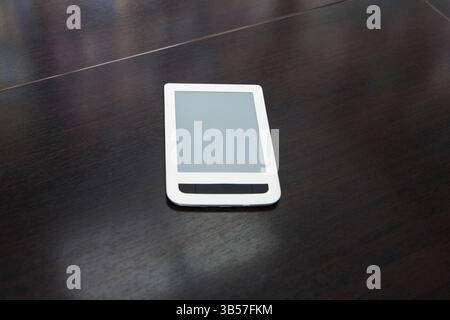 White e-reader with blank screen lying flat on a dark wood table, realistic photo style, minimal office background, concept of digital reading Stock Photo