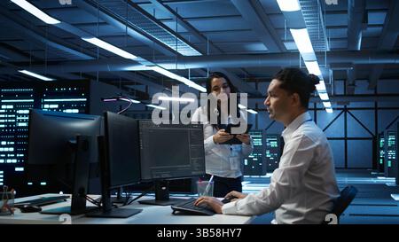 Team of programmers in high tech data center troubleshooting issues using tablet. Teamworking colleagues in server farm using device, establishing performance benchmarks, camera B Stock Photo