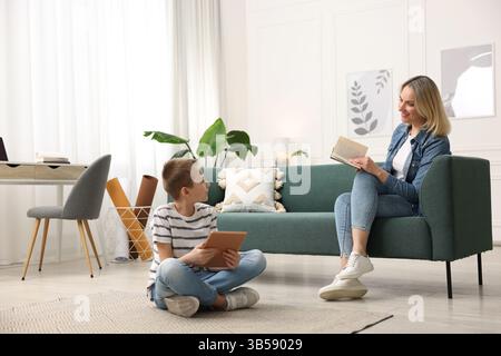 Son using tablet while his mother reading book at home Stock Photo