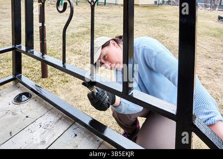 Applying protective coating to metal porch railing, mature white woman in her 50s paints railings with paintbrush. Stock Photo