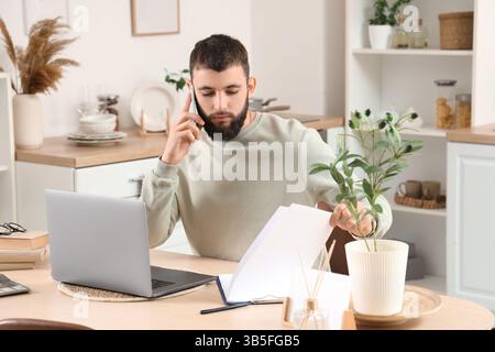 Working young bearded man with clipboard talking by mobile phone at table in kitchen Stock Photo