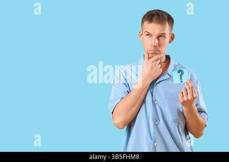 Thoughtful young man holding sticky note with question mark on blue background Stock Photo