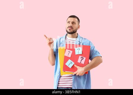 Portrait of thoughtful young man holding folders with question marks on pink background Stock Photo