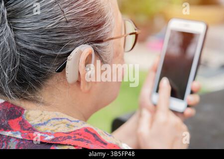 Hearing aids, deafness treatment, innovative technologies at audiology. Mature woman with hearing aid using smartphone at outdoor. Stock Photo
