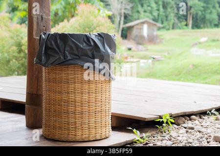 A bamboo wooden trash can stands on the wooden floor in a green park. The trash can is made of woven bamboo. Copy space, selective focus. Stock Photo