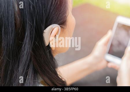 Close up of black hair mature woman wearing hearing aid on ear while using smartphone at home. Selective focus. Stock Photo
