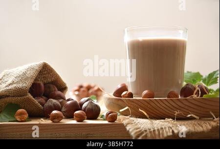 Glass of hazelnut milk on a wooden plate on a wooden table with a sack with hazelnuts and white isolated background. Font view. Stock Photo