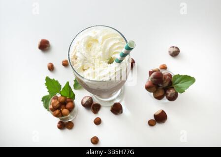 Glass with hazelnut smoothie with cream and green straw on white table with bowls full of hazelnuts and leaves around on isolated on white table. Top Stock Photo