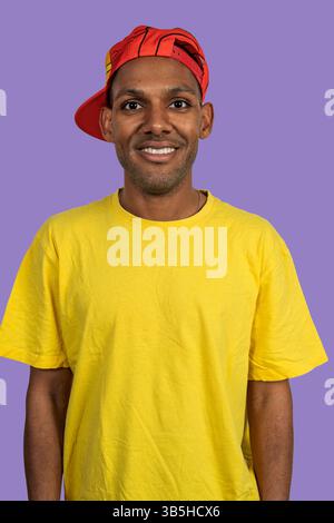 Studio portrait of smiling young man wearing yellow t-shirt and red baseball cap against purple background Stock Photo
