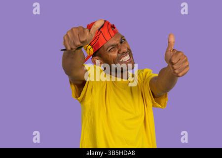 Young man wearing yellow t-shirt and red baseball cap showing thumbs up and pointing finger gun gesture, expressing excitement and approval on purple Stock Photo