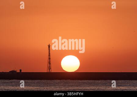 A majestic sunrise unfolds at the beach, with a pier, the sun emerging over a grand sphere, an orange sky, silhouetted birds, and a metallic beacon, c Stock Photo
