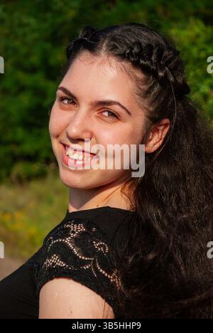 Young caucasian female with braided hair smiling outdoors in sunlit green nature setting, wearing a black lace top and embracing joyful moments. Stock Photo