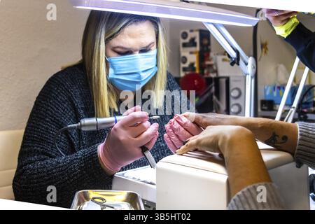 A nail technician wearing a mask works on a client's nails using professional salon tools Stock Photo