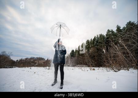 A joyful woman playfully interacts with falling snow, using a transparent umbrella. The snowy scene features a wintery forest in the background. Stock Photo