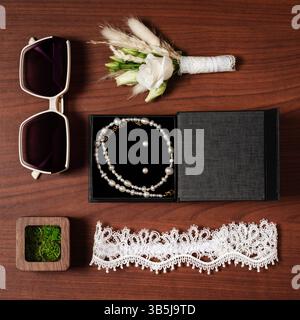 Flatlay of wedding essentials: pearl necklace in a box, earrings, a small bouquet, sunglasses, and a delicate garter. Perfect for bridal preparations. Stock Photo