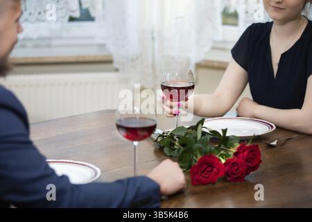 There is a bouquet of red roses on a wooden table. Next to them are empty plates and two glasses of red wine. You can see fragments of the silhouettes Stock Photo