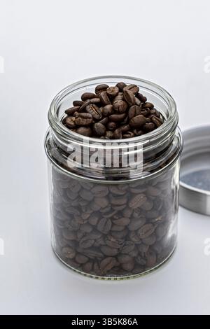 A glass jar filled with aromatic coffee beans, with metal lid placed beside it on white background. image captures rich texture and dark color of bean Stock Photo