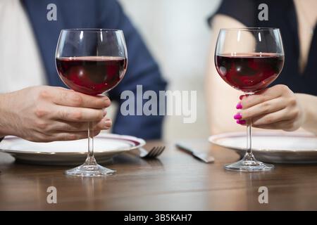 Two glasses of red wine stand on the table. A man and a woman are holding their glasses. In the background you can see blurred figures of a man and a Stock Photo