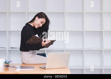 Asian businesswoman sitting on the edge of desk and working in modern office. Stock Photo