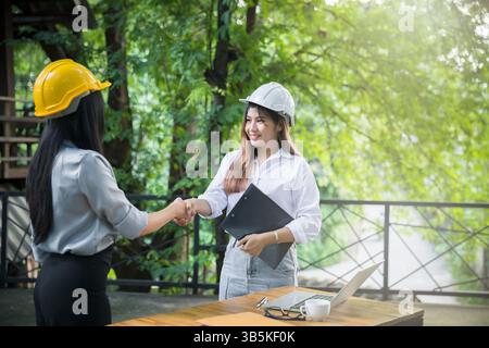 Two asian female architect and engineer in protective helmets shaking hands after meeting. Success collaboration concept. Stock Photo