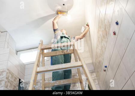 A construction worker applies leveling compound to the ceiling with a stainless steel trowel. A builder dressed in protective clothing is standing on Stock Photo