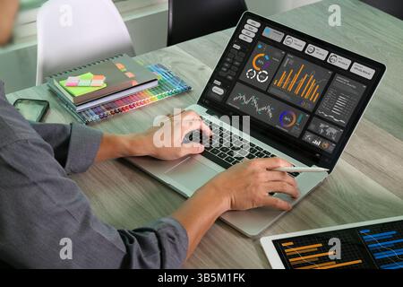 A designer works on a laptop displaying colorful analytics dashboards, with notebooks and pens in the background, representing creative marketing work Stock Photo