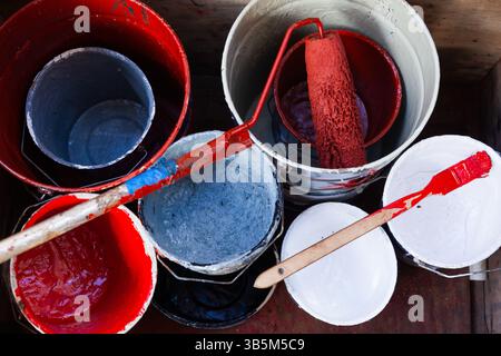 Empty buckets of paint of different colors and a roller and a brush stained with red paint, top view. Stock Photo