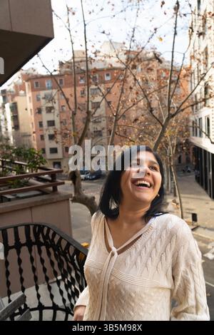A woman is smiling and wearing a white sweater. She is standing on a balcony and looking out at the city Stock Photo