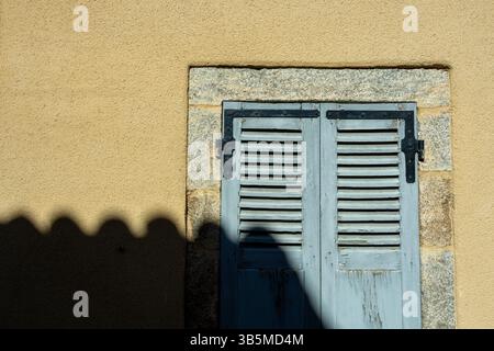 A pair of classic wooden shutters in a soft blue hue sits against a light tan wall. The stone frame adds character with shadows Stock Photo