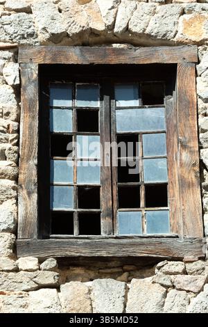 A weathered wooden window with several broken glass panes is set in a rustic stone wall. The sunlight casts shadows, highlighting the textures of the Stock Photo