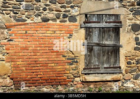 A rustic wall combines textured red bricks and aged stones, complemented by wooden shutters. Sunlight casts shadows. France Stock Photo