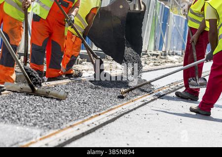 Team of workers put the hot asphalt on a street along tram car's railroad lines. Road construction workers with shovels in protective uniforms and an Stock Photo