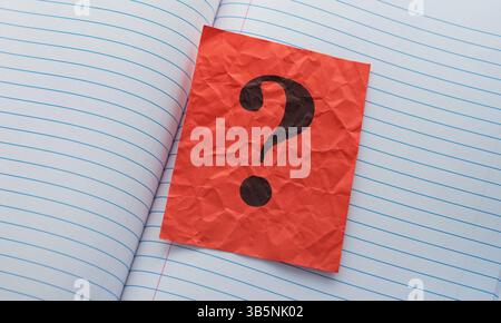A crumpled red paper note with a question mark on it lying in an open notebook. Close up. Stock Photo